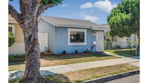 A single-story residential house with a light blue exterior, a front door, and a small front yard with a tree and a sidewalk.
