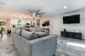 A modern living room with a grey sofa, a ceiling fan, a flat-screen TV, and a kitchen in the background. - Modern Pool Home Near UF, Downtown, & Devil's Millhopper Geological State Park (Gainesville)