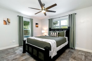 A bedroom with a bed, a ceiling fan, a nightstand, a lamp, and a window with curtains. - Modern Pool Home Near UF, Downtown, & Devil's Millhopper Geological State Park (Gainesville)