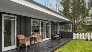 Une véranda couverte avec des fauteuils en osier et une petite table, une clôture blanche en bois et une vue sur la verdure environnante.