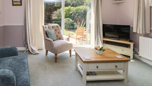Un salon comprenant un canapé, un fauteuil et un téléviseur posé sur un meuble. Une porte-fenêtre coulissante donne accès à un jardin.