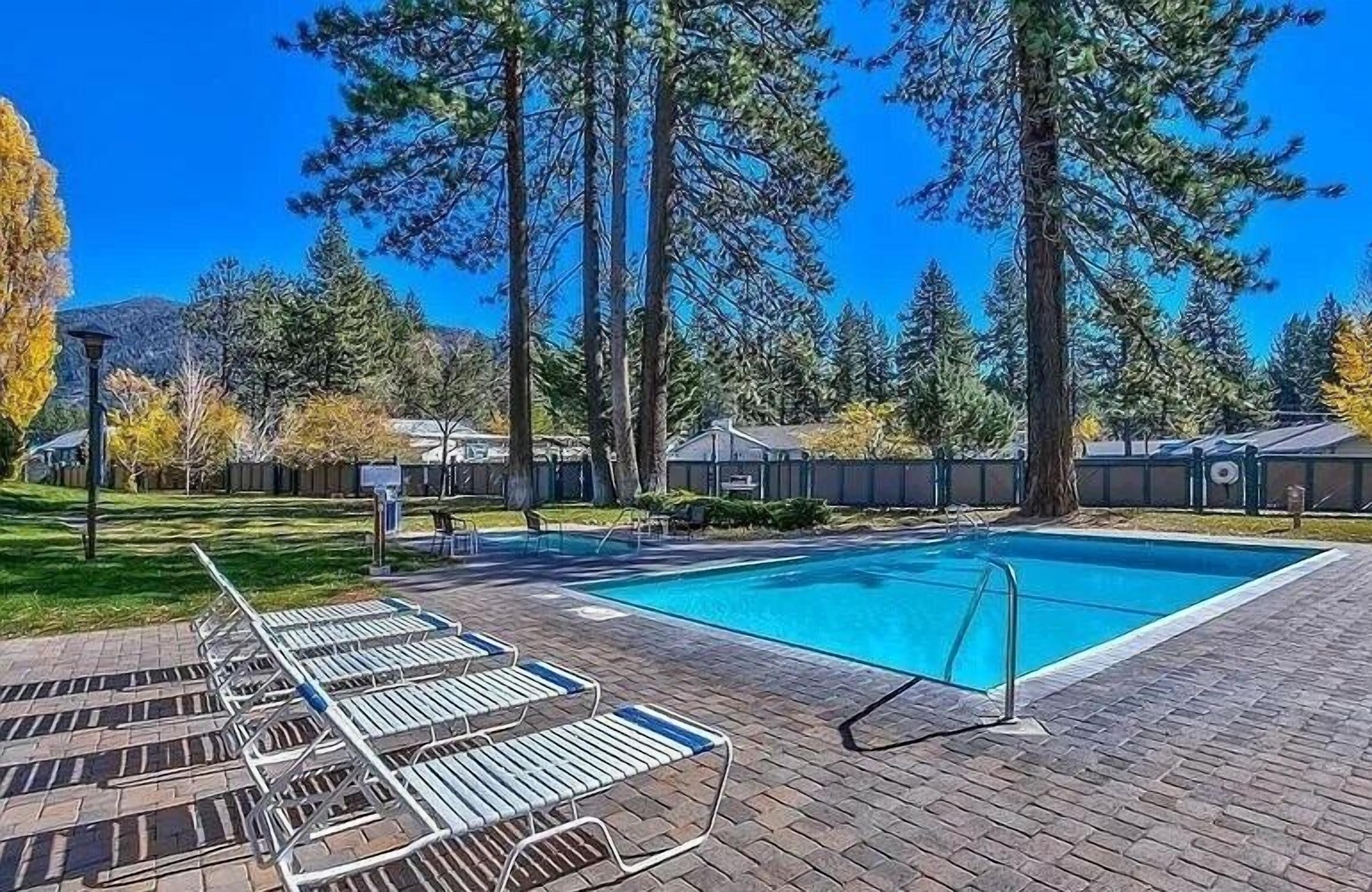 A rectangular swimming pool with a clear blue water surface, surrounded by a paved deck with lounge chairs. Tall trees and a mountain are visible in the background.