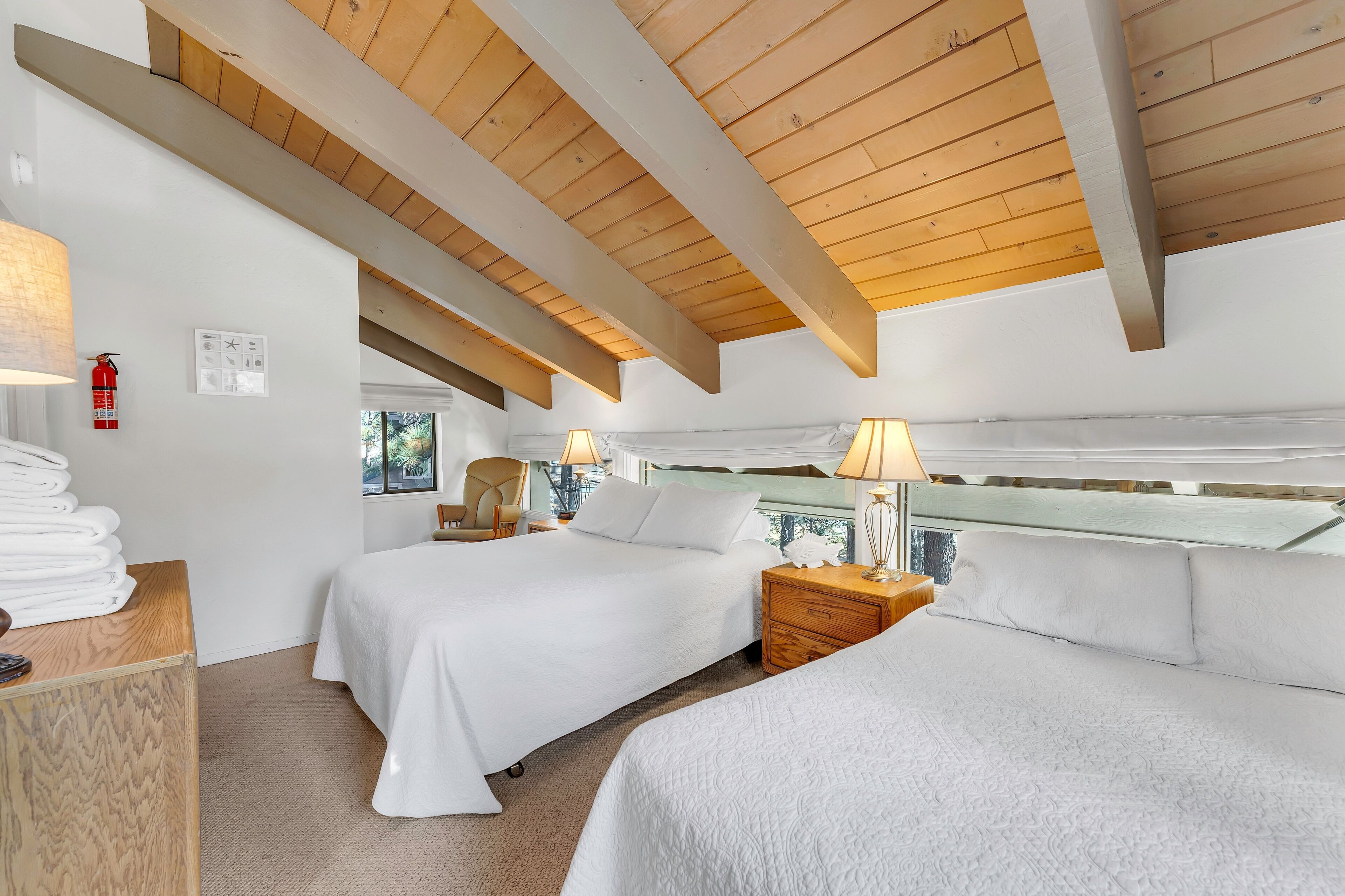 A hotel room with two beds, wooden beams on the ceiling, a bedside table with a lamp, and a window with a view.
