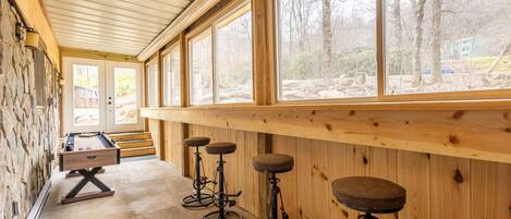 Un espace bar avec des tabourets en bois et une table de billard, de grandes fenêtres offrant une vue sur les arbres et un bâtiment à l’extérieur.