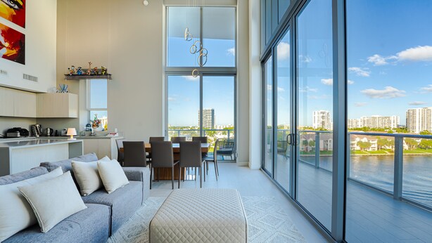A modern living room with a grey sofa, ottoman, and dining area with chairs and a table. Large windows offer a view of a river and cityscape.