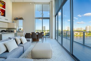 A modern living room with a grey sofa, ottoman, and dining area with chairs and a table. Large windows offer a view of a river and cityscape.