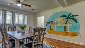 Uma área de refeições com mesa e cadeiras de madeira, ventilador de teto e um mural na parede com uma cena de praia.