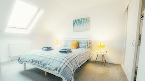 Een slaapkamer met dakraam, een bed met ruitjesdeken, twee bedlampen en een schilderij aan de muur.