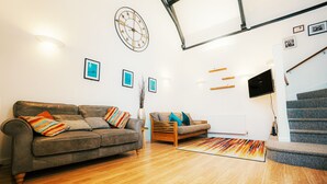 Ein modernes Wohnzimmer mit einem grauen Sofa, einem kleineren Sofa und einem farbenfrohen Teppich. An der Wand hängt eine Uhr, und über einer Treppe ist ein Flachbildfernseher angebracht.