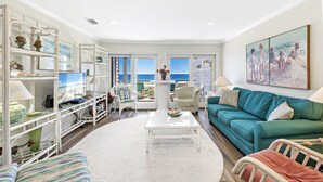 A living room with a view of the ocean, a teal sofa, a white coffee table, and a white bookshelf. - Needle Rush Point D33 | Walk to FloraBama | 2 Pools | Boat Slips | My Beach Getaways (Pensacola)
