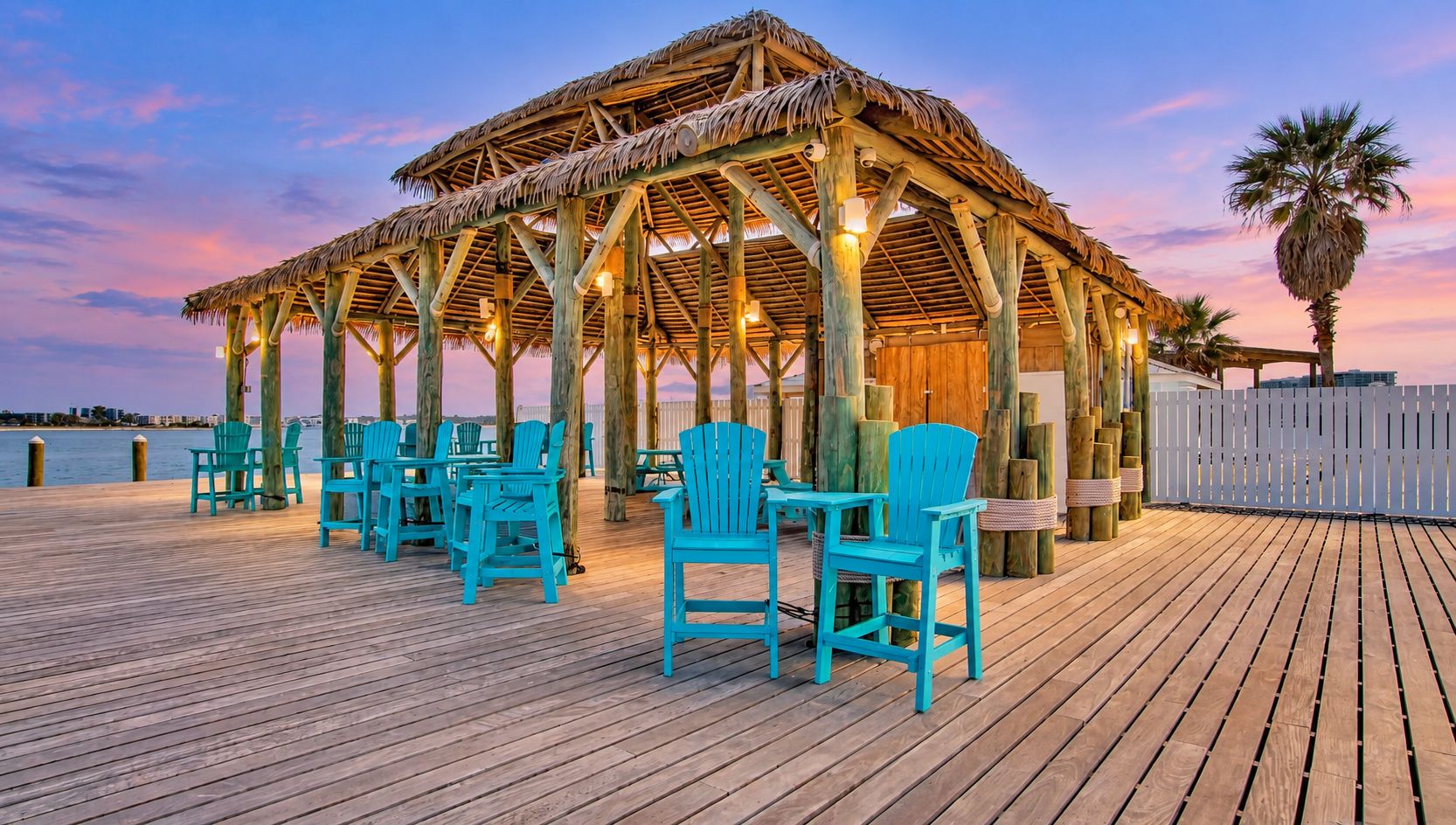 A wooden deck with a thatched-roof gazebo and turquoise chairs.