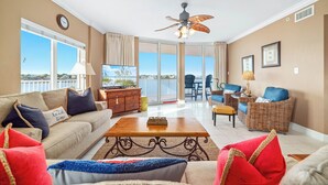 A spacious living room with a sofa, coffee table, and wicker chairs. A television is mounted on the wall, and there is a balcony with a view. - The Pass 301 | Spectacular Views of Perdido Pass | Indoor/Outdoor Pools | My Beach Getaways (Orange Beach)