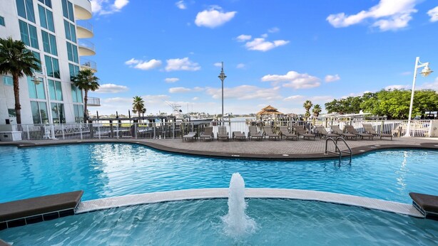 A swimming pool with a fountain, surrounded by a white fence, with a modern building and palm trees in the background. - The Pass 301 | Spectacular Views of Perdido Pass | Indoor/Outdoor Pools | My Beach Getaways (Orange Beach)