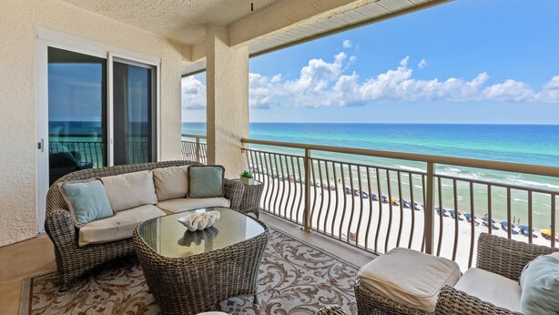 A balcony with a wicker sofa, a glass table, and a view of the beach and ocean.