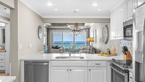 A modern kitchen with white cabinetry, a dishwasher, a stove, and a view of the ocean through large windows.