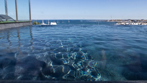 Vista nítida de uma piscina infinita com borda de vidro, que se estende até uma marina com barcos e uma linha costeira distante.