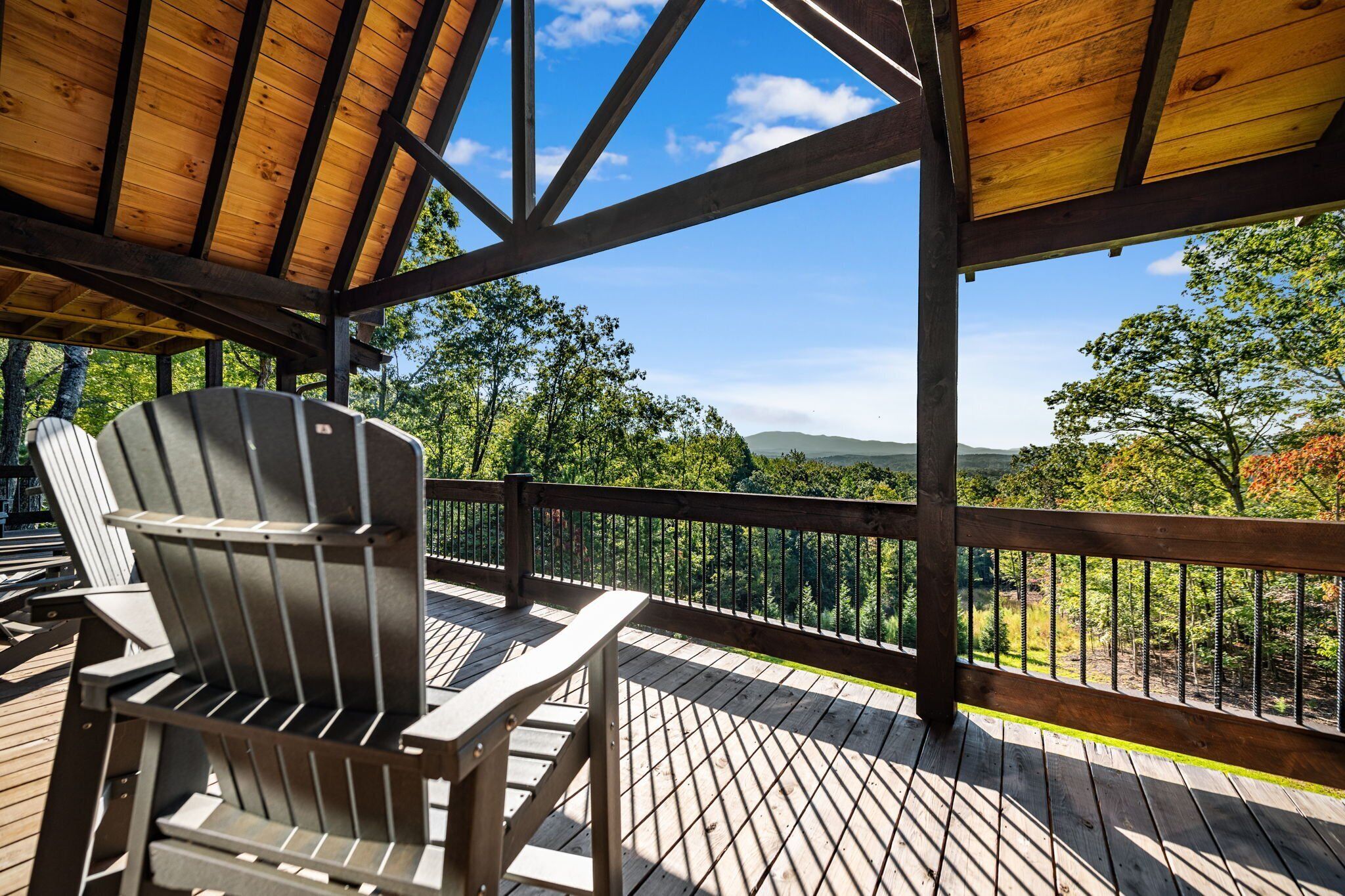 A wooden deck with Adirondack chairs, a wooden railing, and a view of trees and hills.