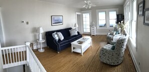 Un salon avec un canapé bleu foncé, une table basse blanche et un fauteuil à motifs. On y trouve une balustrade blanche, un ventilateur de plafond et une grande fenêtre donnant sur un beau paysage.