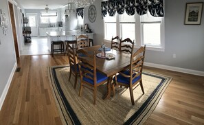 Un coin repas avec une table et des chaises en bois, une cuisine à l’arrière-plan et une grande fenêtre avec des rideaux.