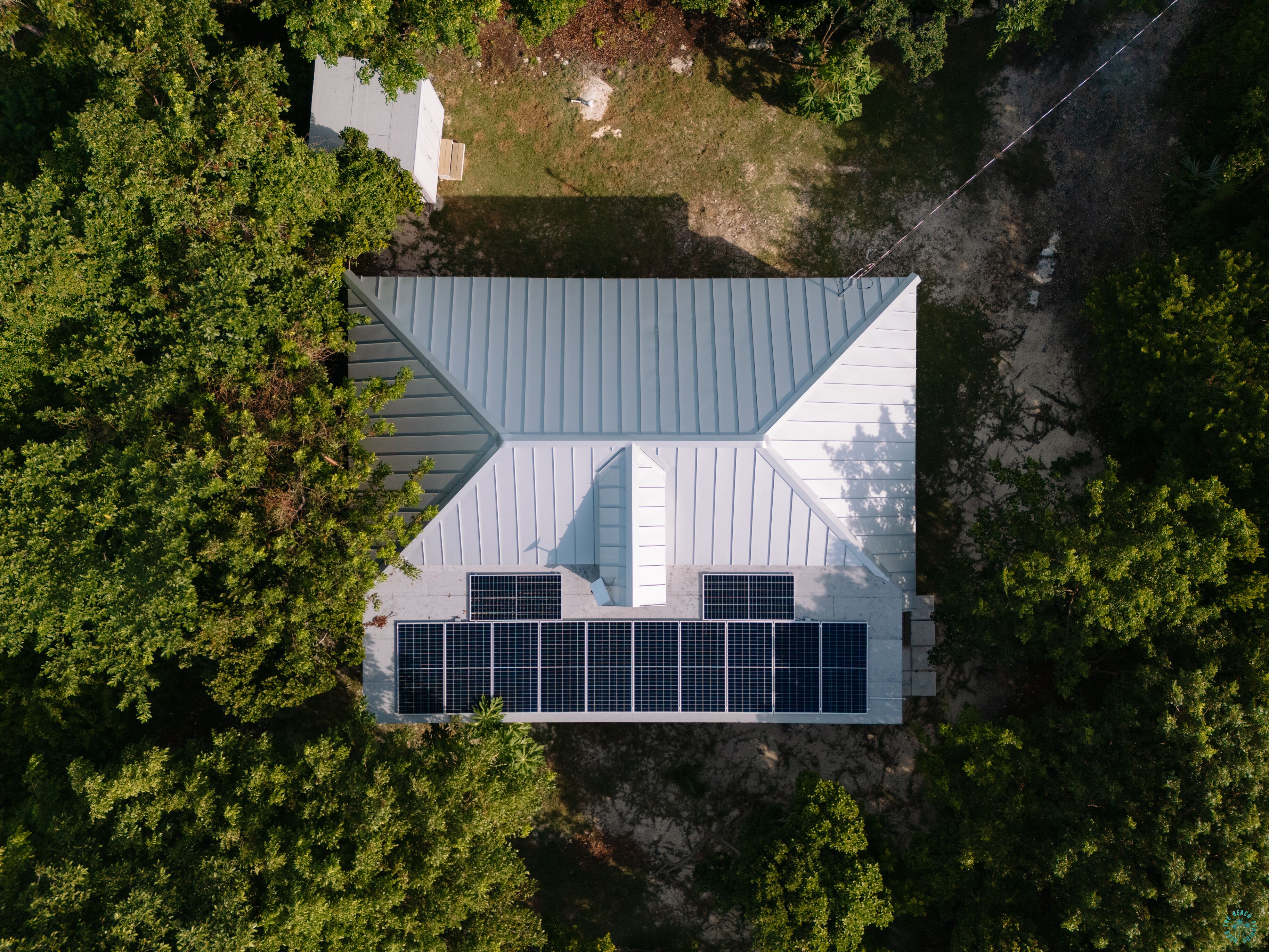 An aerial view of a house with a sloped roof, solar panels, and surrounded by dense trees.