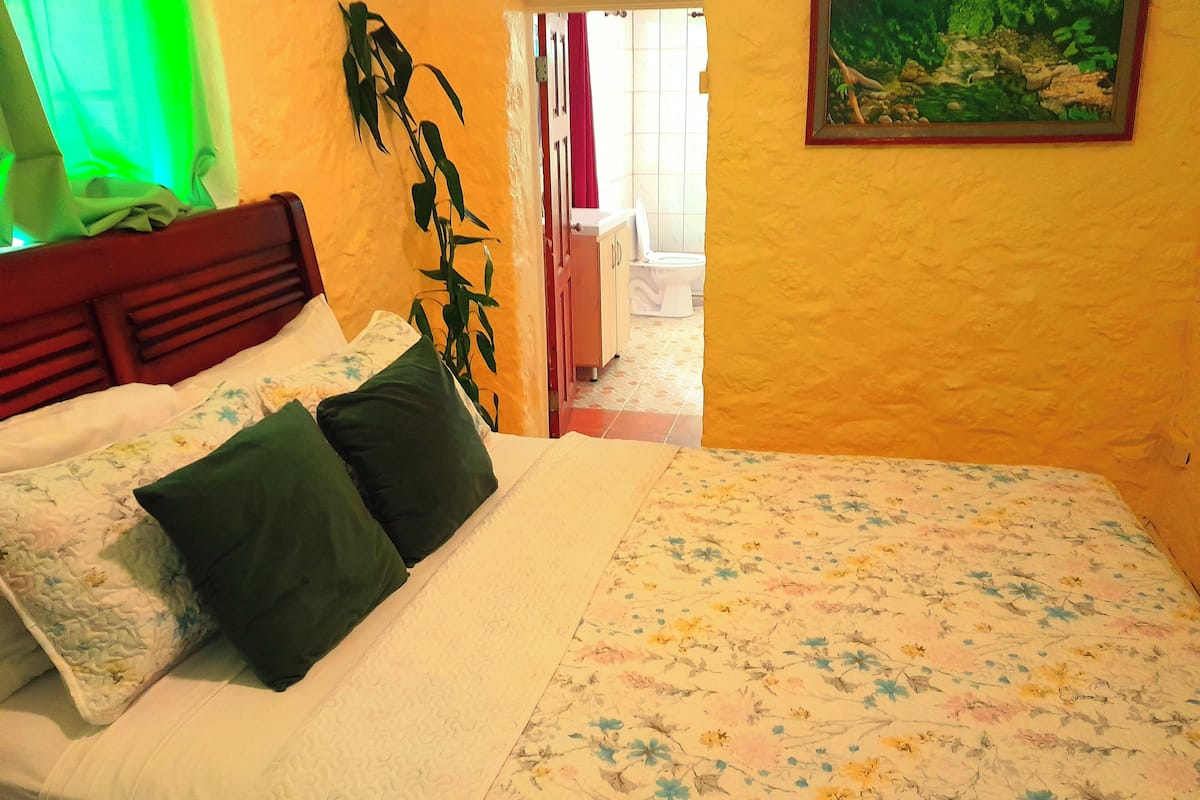 A bedroom with a bed, pillows, a plant, and a painting on the wall.