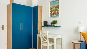 Una habitación con un escritorio blanco, un armario azul, una silla blanca, una pequeña estantería con una planta y un cuadro en la pared.