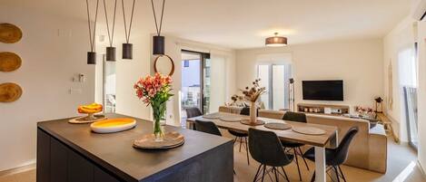Una cocina moderna con una isla central, comedor con sillas y un televisor en la pared.