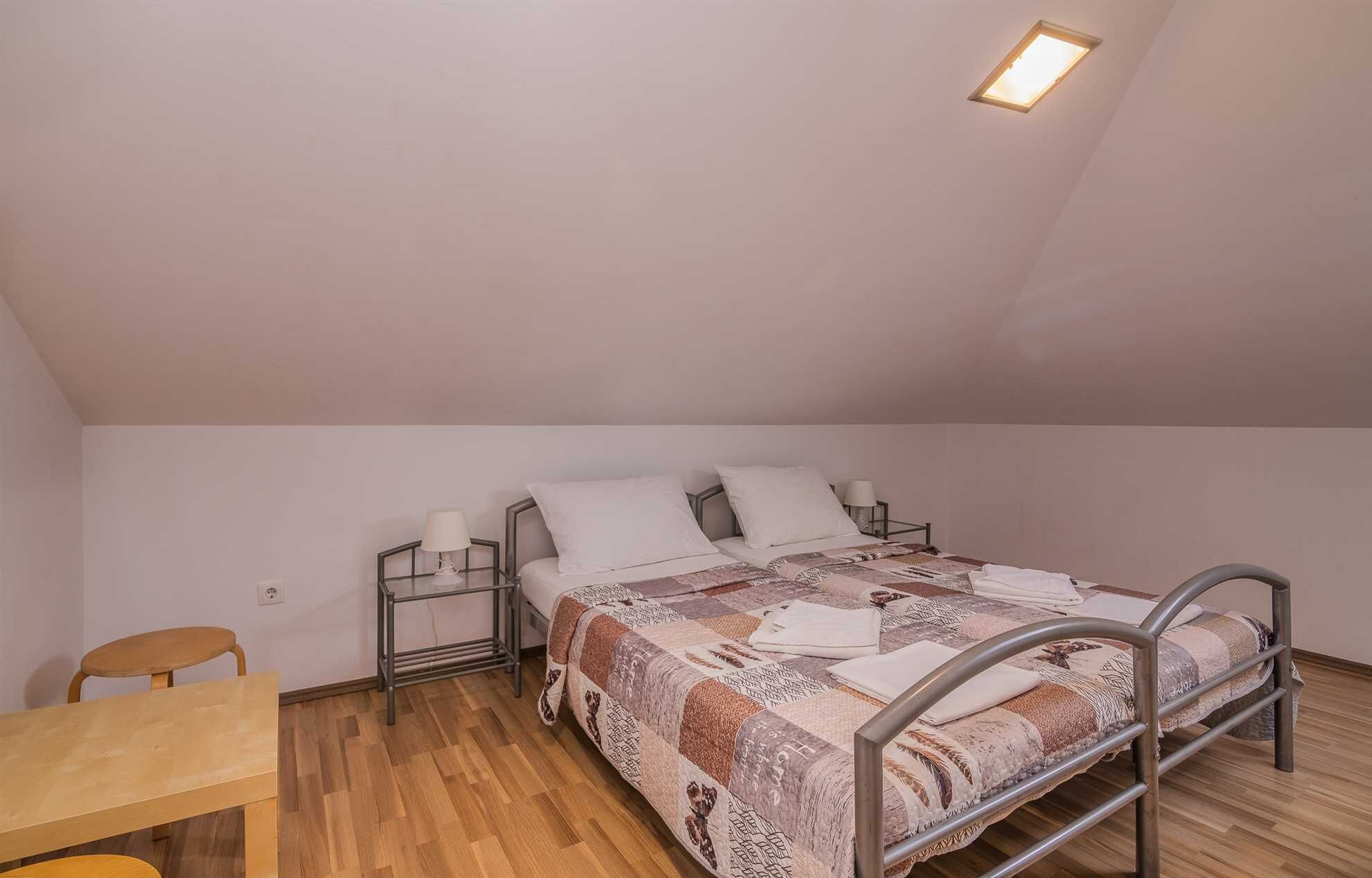 Una habitación pequeña con cama individual, techo inclinado, una silla de madera, mesita de noche con lámpara y una ventana con aplique.