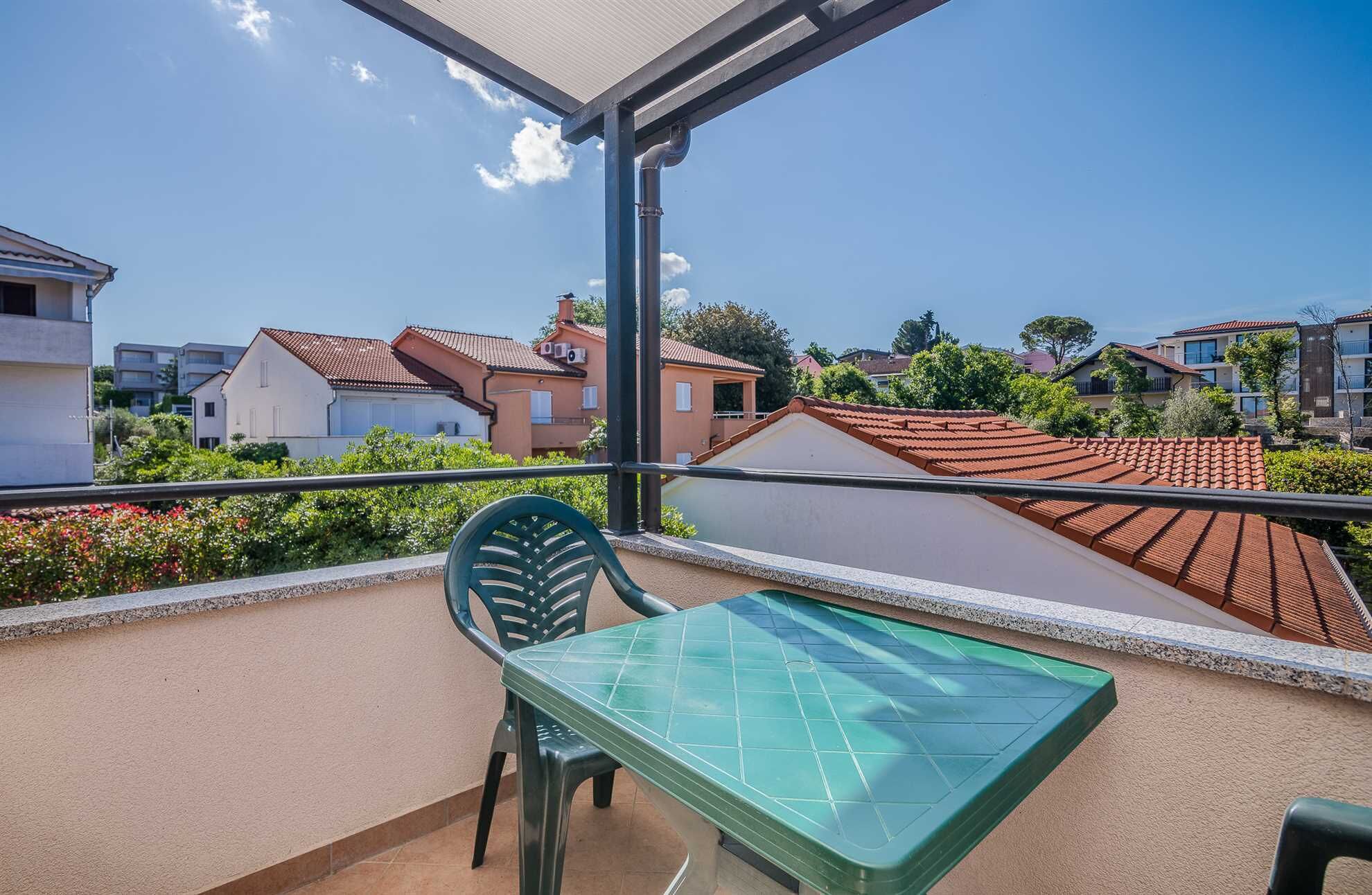 Un balcon avec une table et des chaises vertes, donnant sur un quartier résidentiel avec des maisons et de la verdure.
