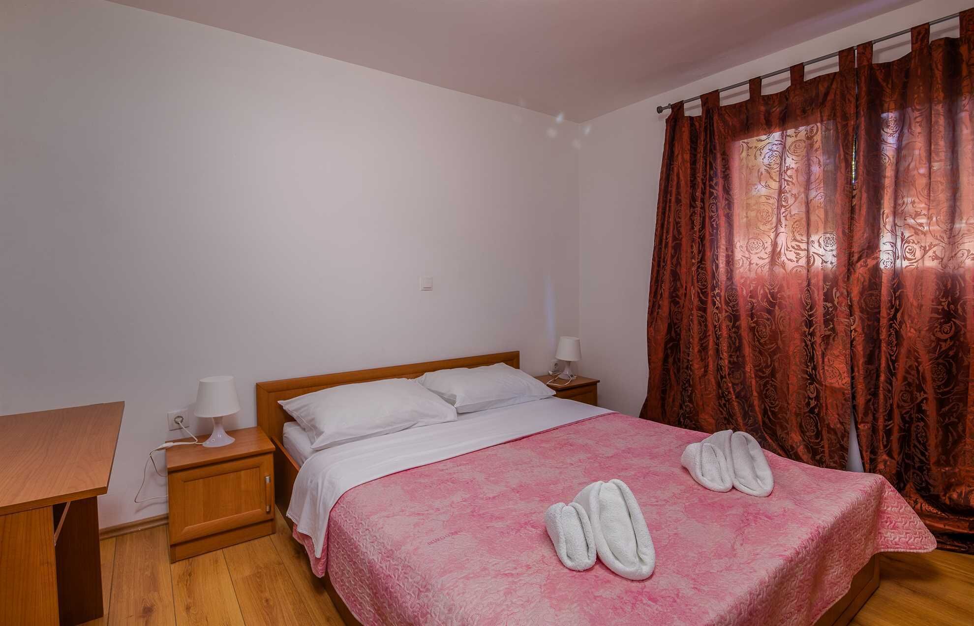 A bedroom with a wooden bed, white bedding, a pink blanket, two white towels, a nightstand with a lamp, and a window with patterned curtains.