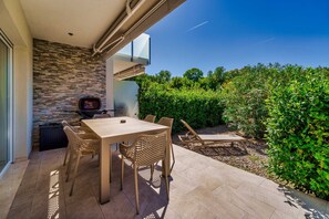 An outdoor patio with a table and chairs, a grill, and a view of a garden.
