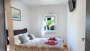 Un dormitorio con cama, almohadas, una ventana y vista a una palmera, y una puerta.