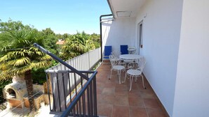 Un balcón con mesa y sillas blancas, una pequeña parrilla y vistas a zonas verdes.