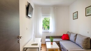 Un espacio habitable compacto con un sofá, una mesa de centro, un televisor montado en la pared y una ventana con cortinas translúcidas.