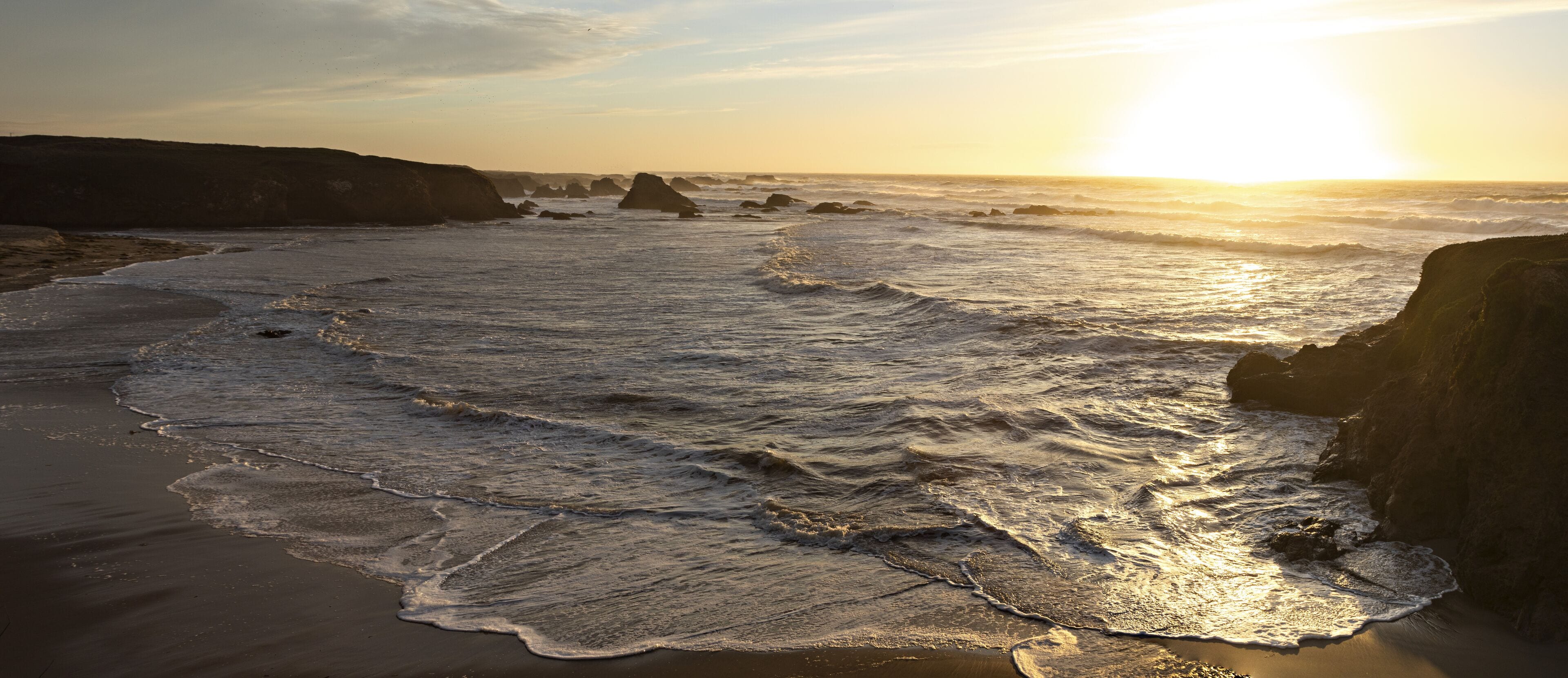 A beach with waves, rocky formations, and the sun setting or rising.