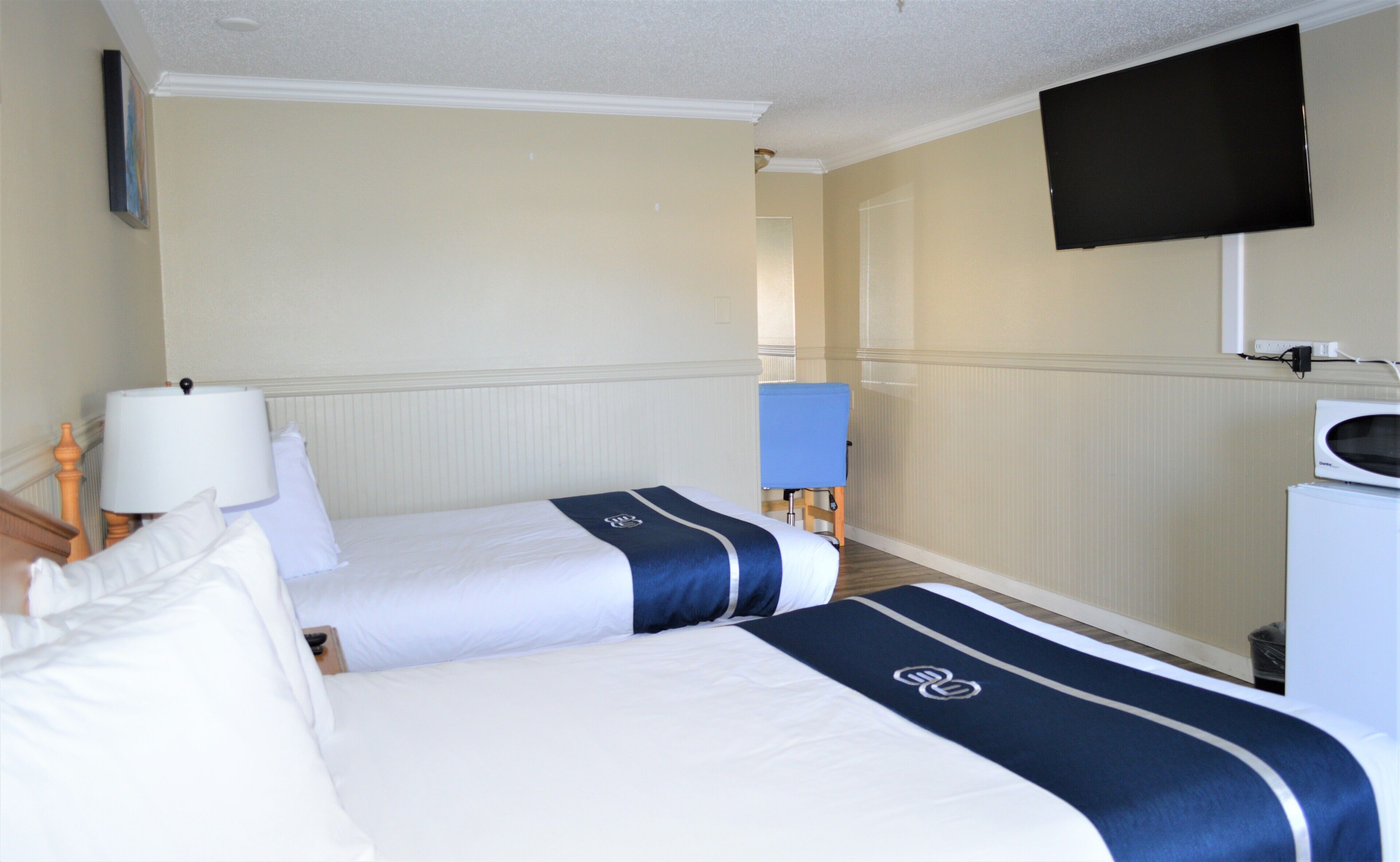 A hotel room with two beds, a TV mounted on the wall, a desk with a lamp, and a microwave.