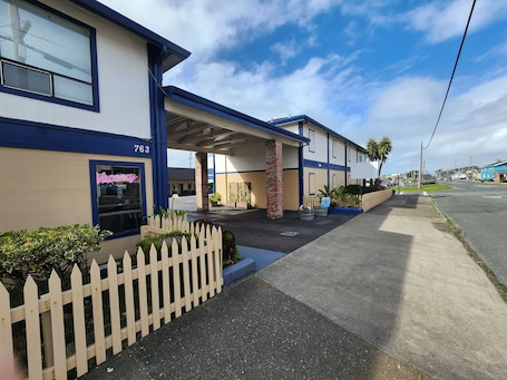 A motel with a blue and beige exterior, a white picket fence, and a neon sign.. Studio 9 Fort Bragg, LLC.