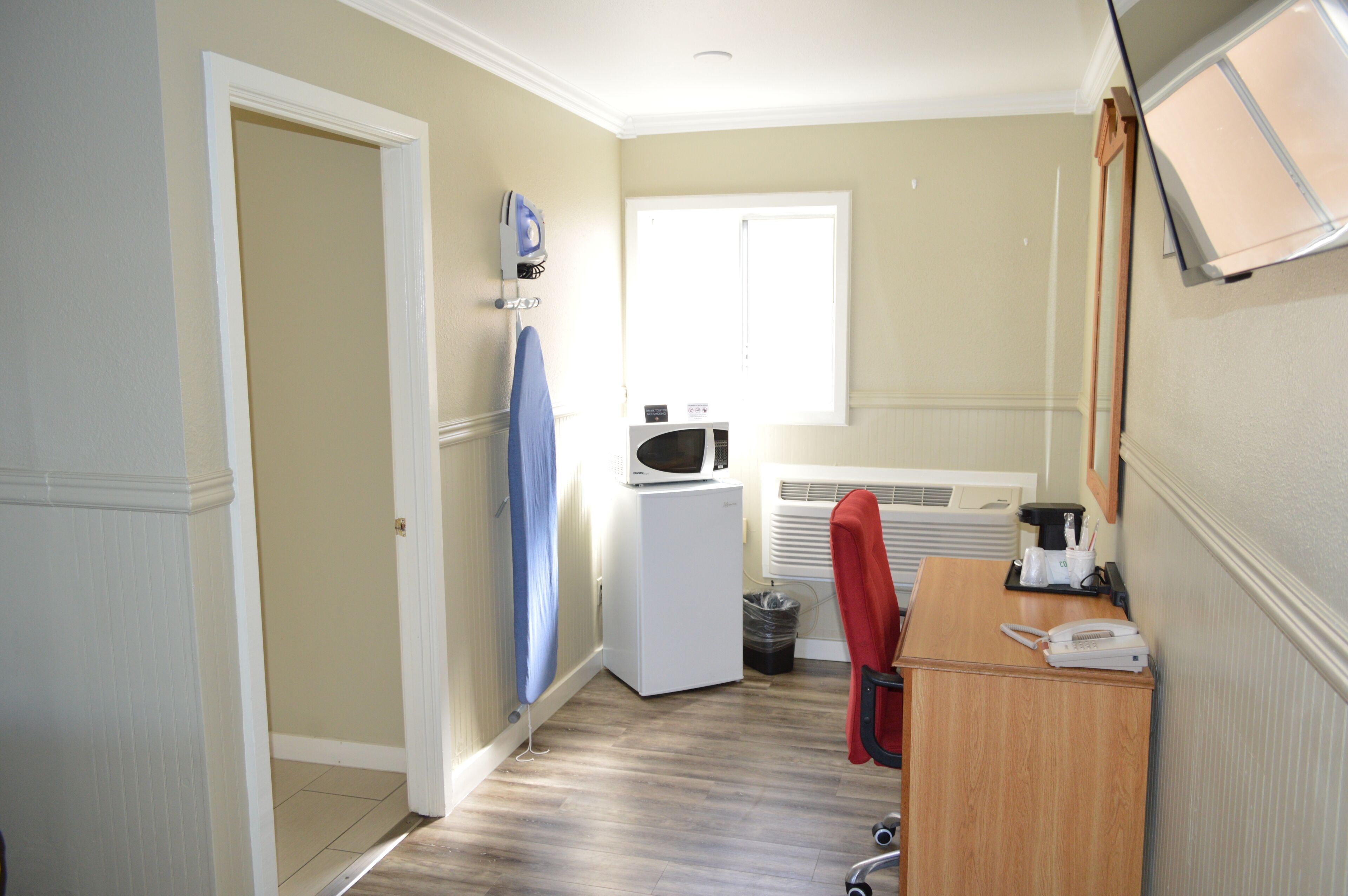 Habitación de hotel pequeña con kitchenette, microondas, refrigerador compacto, escritorio con teléfono y ventana que proporciona luz natural.