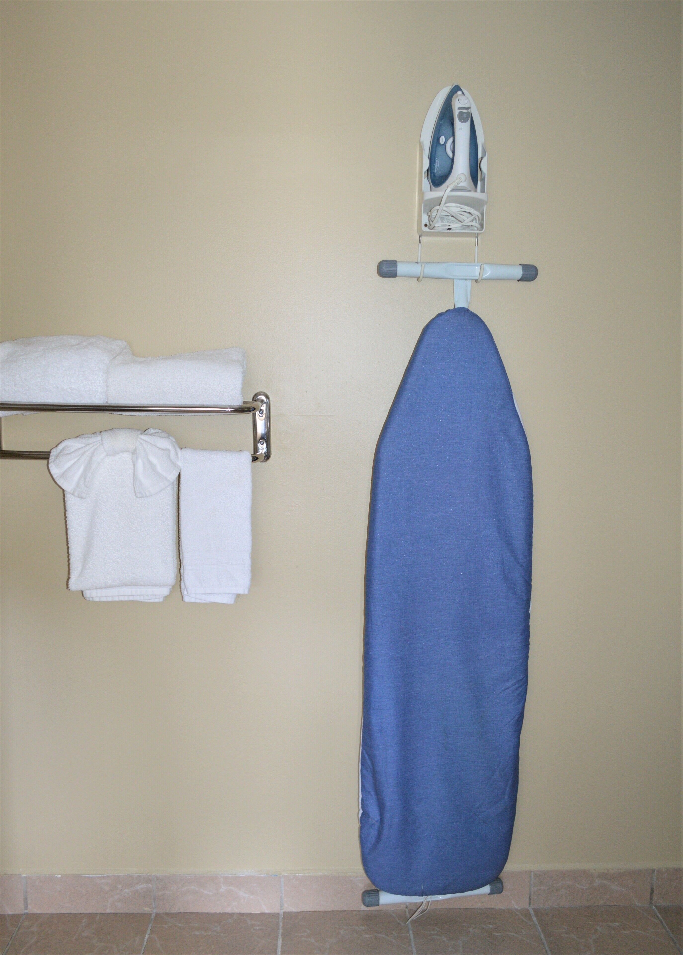 A bathroom with a towel rack holding two white towels, a blue ironing board, and an iron on the wall.