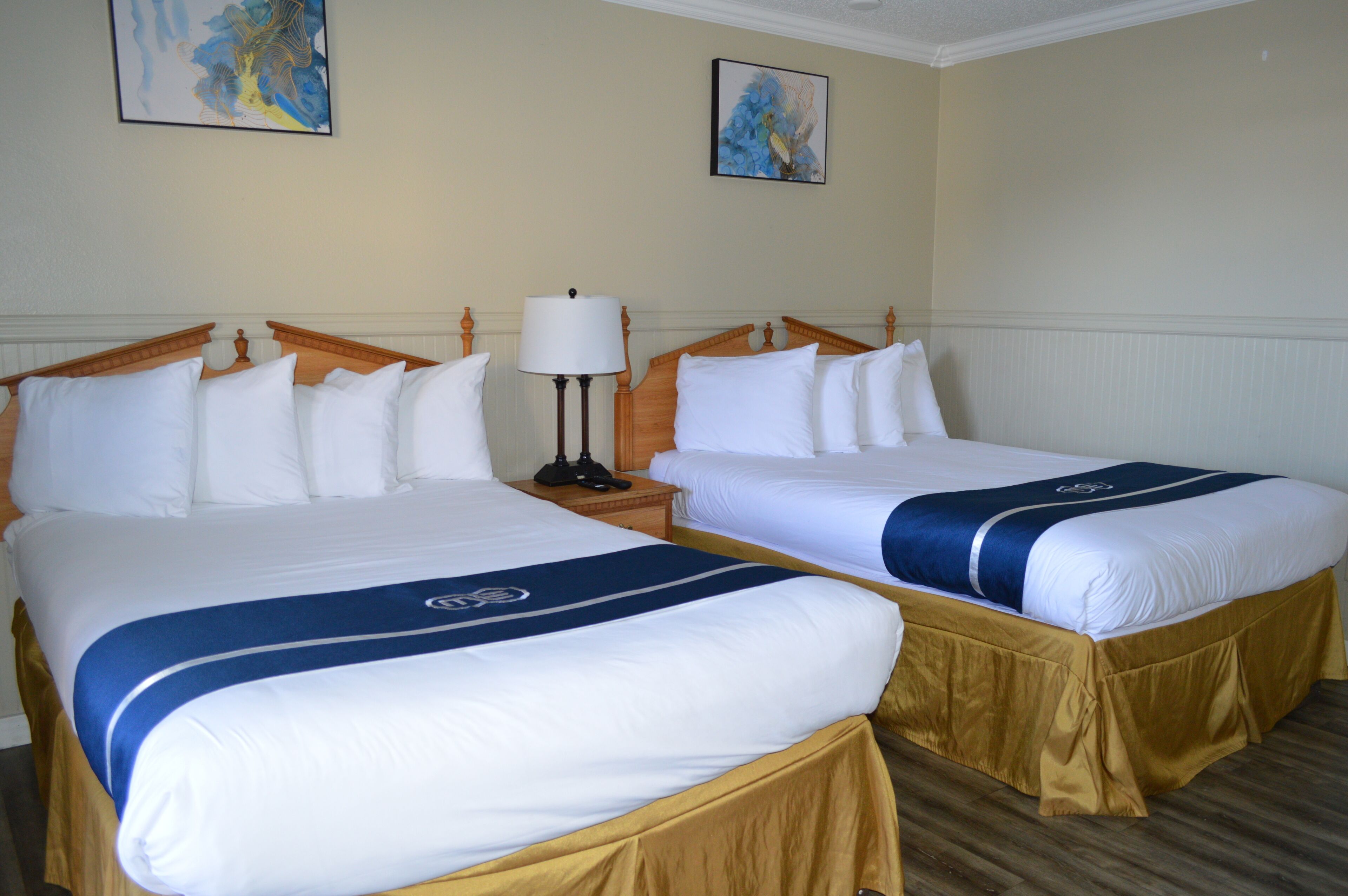 A hotel room with two beds, each with white bedding and a blue accent. There is a nightstand with a lamp between the beds.