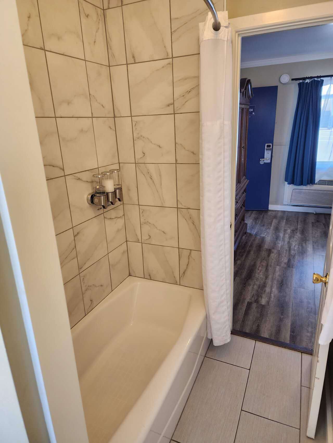 Un baño con bañera, zona de ducha y una puerta que da a un dormitorio con cama y ventana.