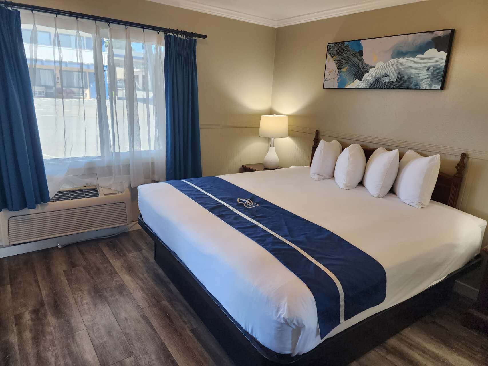 A hotel room with a large bed, a bedside table with a lamp, a window with curtains, and a painting on the wall.