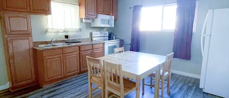 Una cocina con armarios de madera, un refrigerador blanco, una mesa de comedor con sillas y una ventana con cortinas.