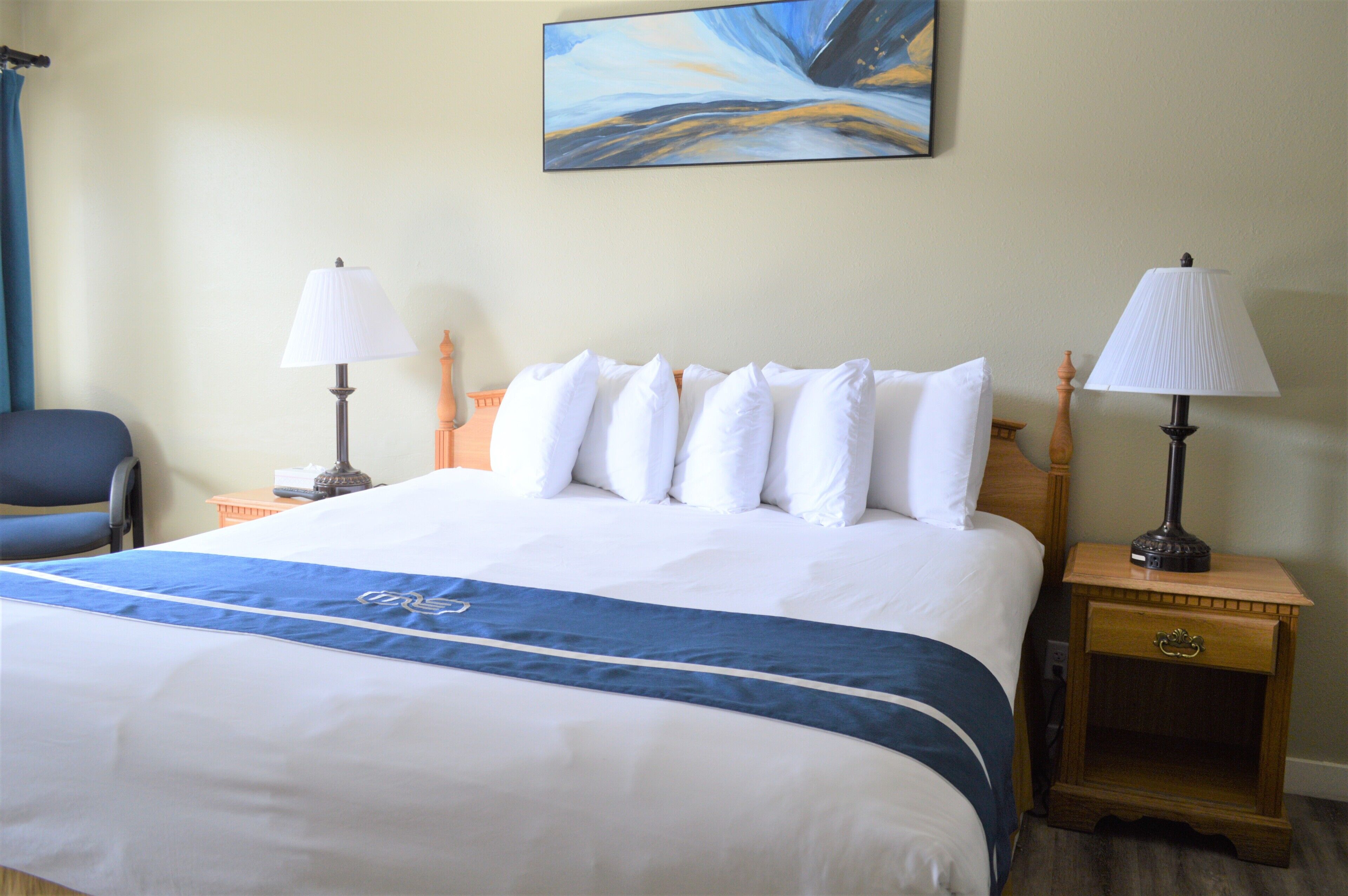 Una habitación de hotel con una cama grande, dos mesitas de noche con lámparas, una silla y un cuadro en la pared.