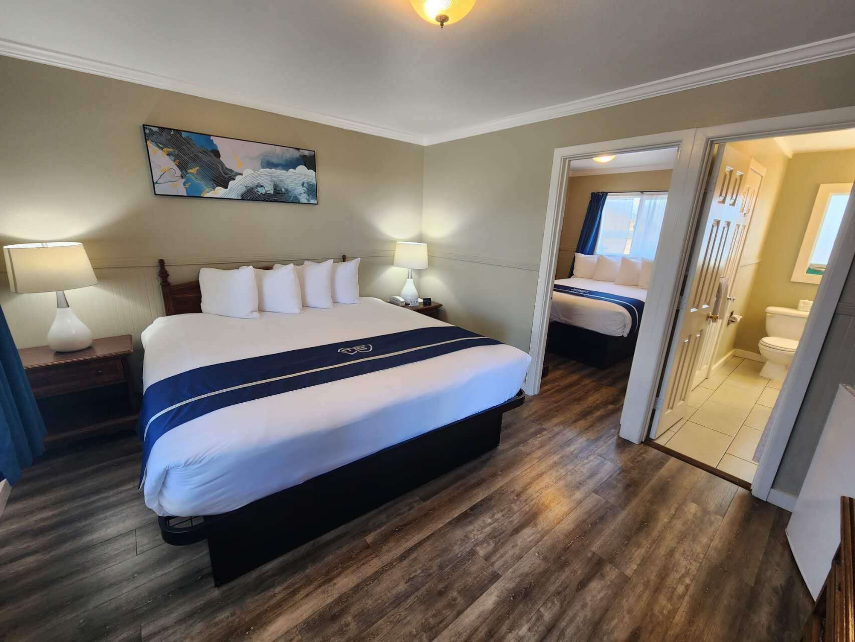 Una habitación de hotel con una cama grande, una mesita de noche, una lámpara, un cuadro en la pared y un baño visible a través de una puerta abierta.