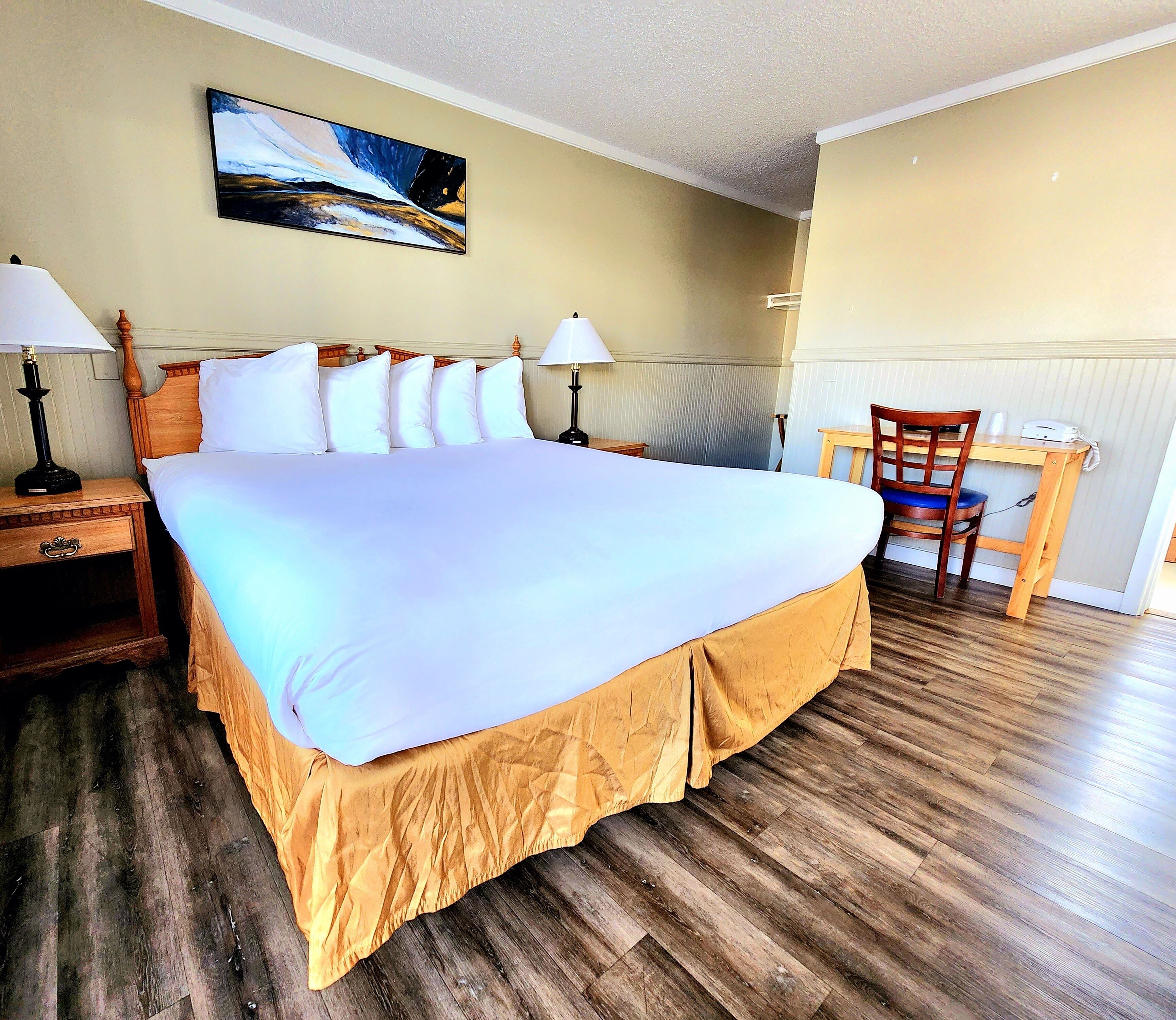 A hotel room with a large bed, two bedside tables with lamps, a wooden desk with a chair, and a painting on the wall.