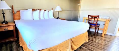 A hotel room with a large bed, two bedside tables with lamps, a wooden desk with a chair, and a painting on the wall.