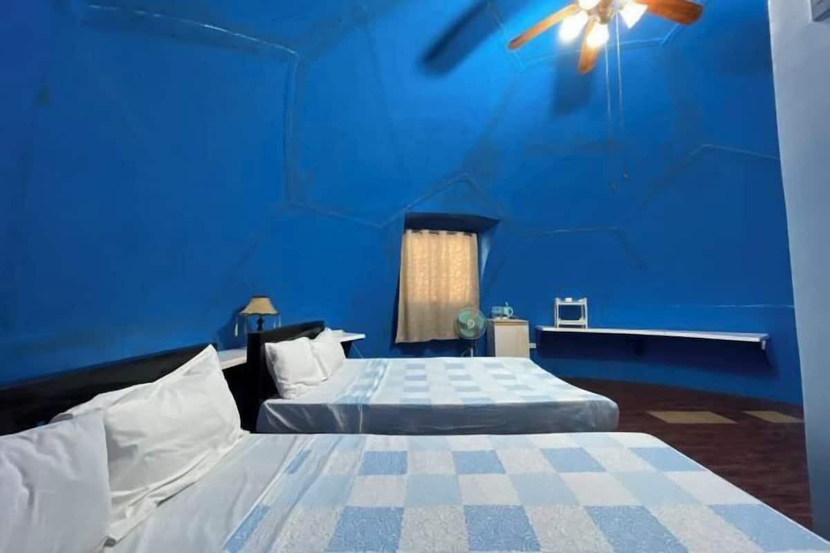 Een kamer met twee bedden, een plafondventilator, een raam met gordijnen, een nachtkastje en een kleine tafel met een lamp.