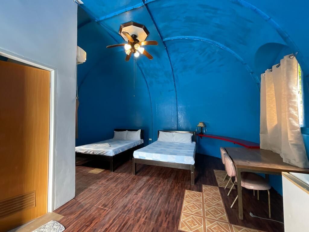 Une chambre de taille modeste, équipée de deux lits simples, d’un bureau avec une chaise, d’un ventilateur de plafond et d’une petite table.