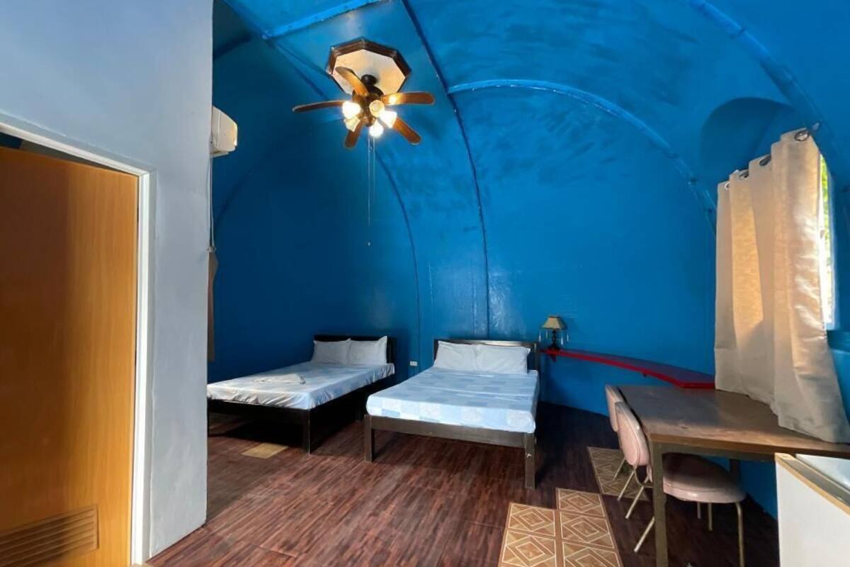 Een compacte kamer met twee enkele bedden, een bureau met stoel, een plafondventilator en een kleine tafel.